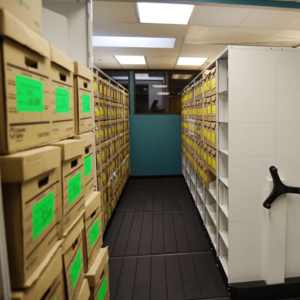 A storage room equipped with Pegasuss RFID Failsafe Medical Inventory System features shelves lined with cardboard boxes labeled bright green on the left and yellow on the right. The black floor contrasts with fluorescent-lit ceilings, and a manual rotating handle is visible on the right shelf.