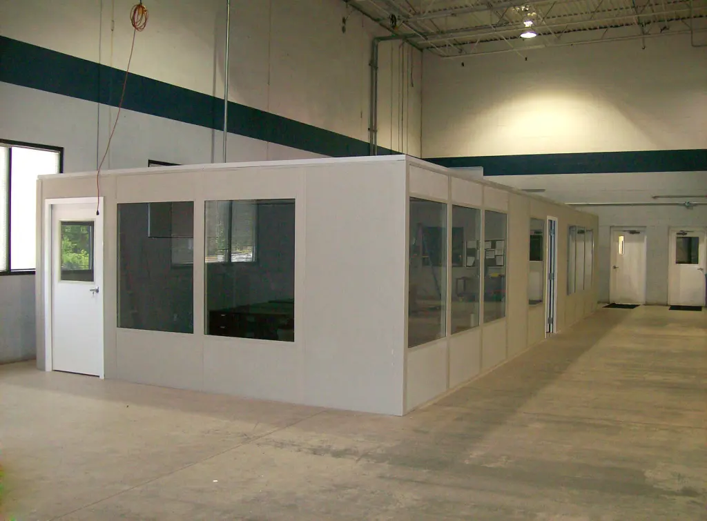 A large indoor space features a simple modular office at its center, ideal for shipping and receiving tasks. The office boasts several windows and a door, while the surrounding warehouse offers high ceilings and industrial lighting.