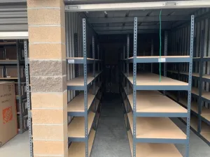 Inside the storage unit, empty metal archive shelving for law firms lines up in two rows, forming narrow aisles. The garage-style door stands open, revealing concrete walls and the edge of a cardboard box on the left.
