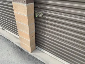 A light brown, corrugated metal storage unit door with a lock on the right side hints at its purpose—perhaps to house archive shelving for law firms. Adjacent to a brick pillar, the setting is complete with an asphalt-paved ground.