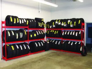The storage room features automotive shelving with two red metal racks, each holding multiple black tires tagged in white. A stark white wall and a bare concrete floor amplify the industrial feel, while overhead fluorescent lights cast their bright glow across the space.