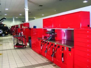 A row of bright red tool cabinets and workstations is neatly arranged with automotive shelving in a car service garage. Tools hang from the workbenches, and various accessories are organized. In the background, a vehicle is being serviced on the white-tiled floor.