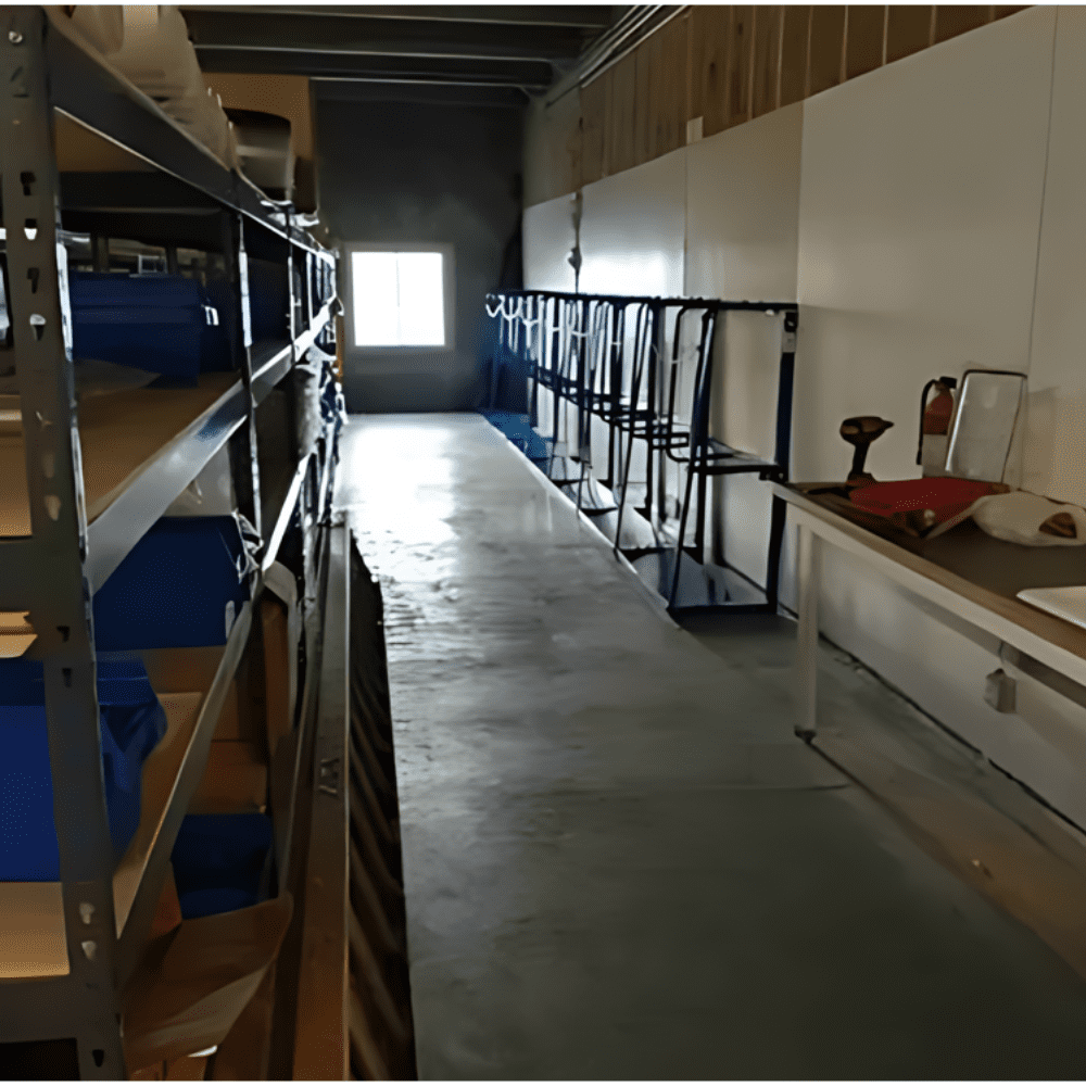 A narrow warehouse aisle features boltless shelving on the left, stacked with blue bins and cardboard boxes. The right side hosts a long table with a fire extinguisher and tools. Sunlight pours in through a window at the far end, illuminating this organized space for a trailer manufacturer.