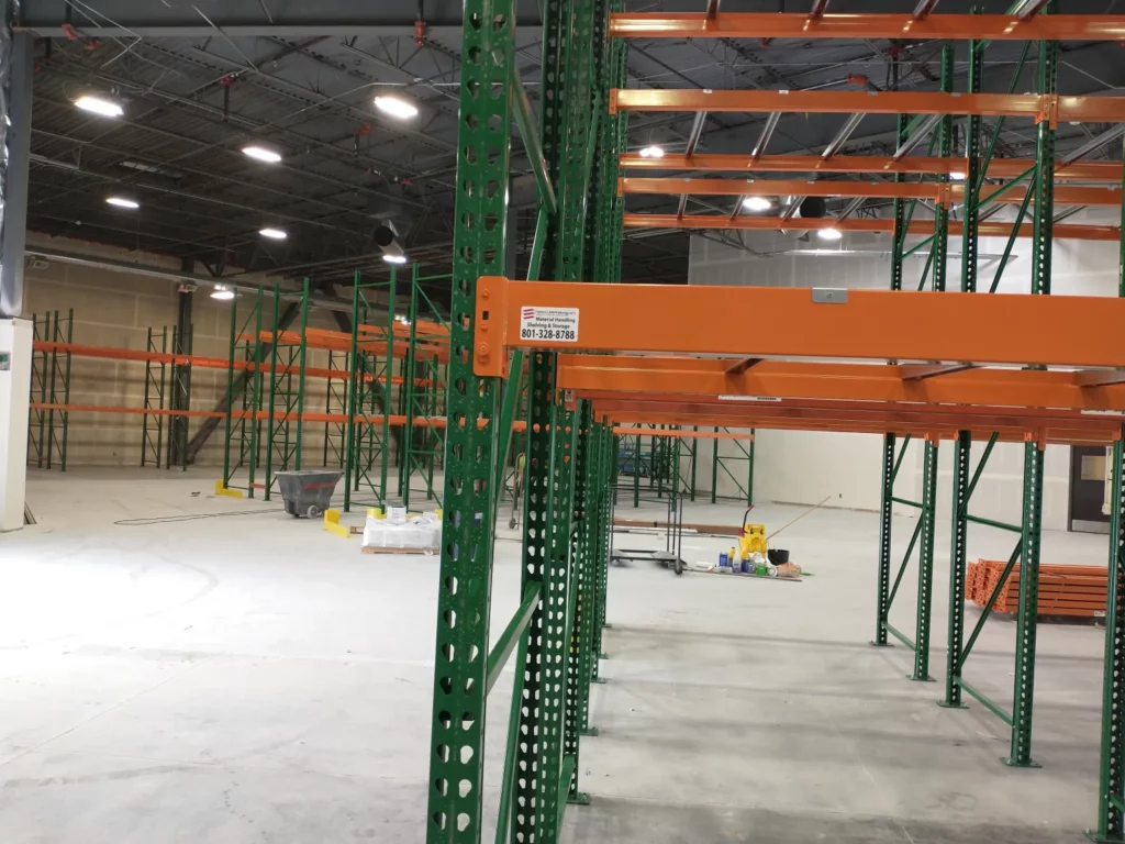 Empty industrial warehouse with green and orange metal shelving racks. The ceiling has exposed beams and lighting fixtures. Some construction materials, including a carpet roll near a pallet rack, are on the floor, indicating ongoing setup or renovation.