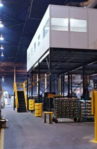 A warehouse interior features a white modular office structure elevated on black metal supports. A yellow staircase leads up to it. The floor is open, with carts and industrial equipment visible, ideal for shipping and receiving. Overhead lighting brightens the space.