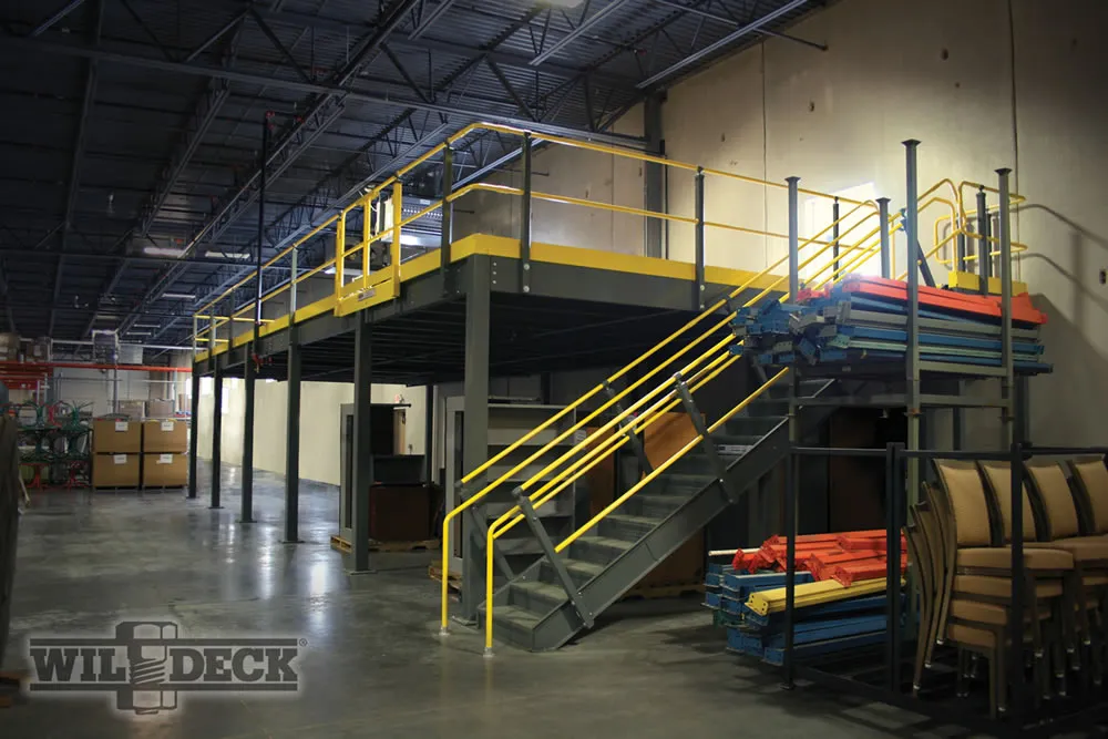 Industrial warehouse interior featuring a gray metal mezzanine with yellow railings and stairs. Stacks of colorful pallet racking materials, ideal for organizing, are nearby. The scene is well-lit with overhead fluorescent lights. The Wildeck logo is in the lower left corner.