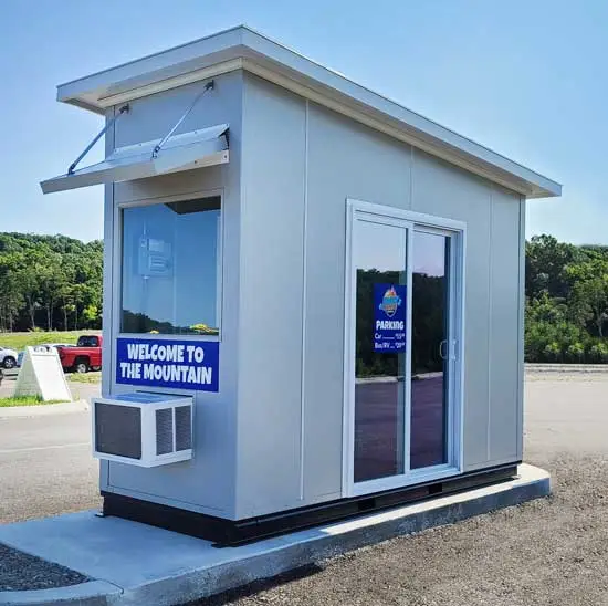 A small, modern booth with large windows, set on a concrete platform, reminiscent of Utah guard booths. A sign reads Welcome to the Mountain, with parking costs detailed on the door. Its nestled in a parking lot, surrounded by trees in the background.