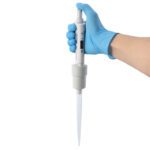 A hand in a blue glove holds an MH-USA Precipette Pipette, single-channel with adjustable volume of 1-10ml, featuring a display and long narrow tip for precise laboratory liquid measurement and transfer.