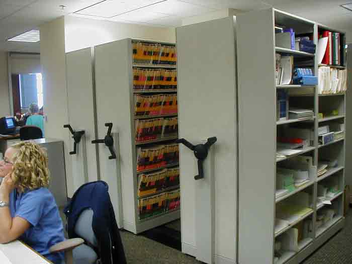 In a well-lit office, rows of mobile shelving units for medical records hold files efficiently. A person with curly hair sits in the foreground, working on a computer. Office supplies and binders are neatly organized on an adjacent shelf, ensuring seamless health information management storage.