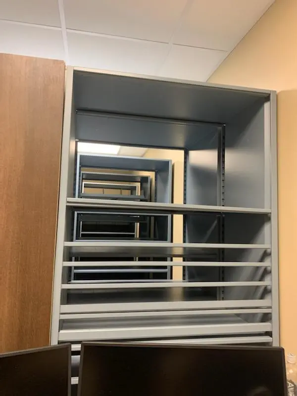 A series of empty metal frames aligned like a tunnel, creating an optical illusion of infinite depth. These mobile shelving units for pharmaceutical supplies are set against a light-colored wall, with part of a wooden door and computer monitors visible in the foreground.