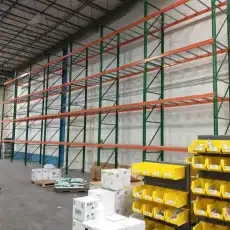 A large warehouse with tall, empty metal storage racks against a wall resembles Utah lockers. In the foreground, yellow storage bins, white boxes, and scattered items are visible on the floor. The ceiling is high with visible beams.