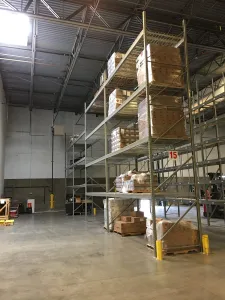 The interior of a warehouse features tall metal shelving units filled with stacked boxes, showcasing efficient pallet rack solutions ideal for auto glass distribution centers. The concrete floor and natural light streaming through a skylight highlight sparse items scattered below.
