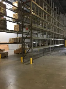 Discover an expansive industrial warehouse featuring tall metal shelving units, ideal for pallet rack solutions in auto glass distribution centers. Shelves hold cardboard boxes and wooden pallets. The concrete floor and plain walls are accented by yellow safety posts securing the base of each shelf.