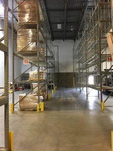 A warehouse interior with tall metal shelves filled with wrapped pallets showcases an ideal pallet rack solution for auto glass distribution centers. The concrete floor and yellow safety barriers ensure safety, while numbered signs 14 and 15 help maintain organization.