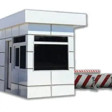 A small, modern-looking, panel-built white guard booth with large windows and a flat roof stands beside a red and white striped barrier gate. The background is plain white.