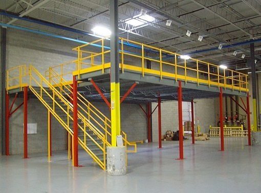 A warehouse mezzanine with a gray floor and bright yellow guardrails, supported by red beams. A matching yellow staircase provides access to the upper level. The area is spacious with a concrete floor and high ceiling.