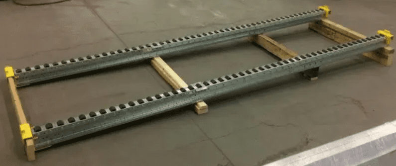 A metal frame with two long parallel beams, supported by wooden blocks at both ends and the center, resembles a pitch-rail pallet rack. The beams have evenly spaced rectangular notches along their lengths. The setup is firmly arranged on a concrete floor.