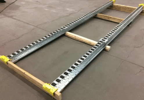 Two parallel metal tracks, reminiscent of a pitch-rail pushback pallet rack, are connected by wooden beams on a concrete floor, forming a rectangular frame with yellow brackets at the ends. The setup resembles a simple, low-level ladder or support structure.