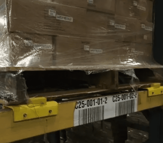 Stacks of cardboard boxes wrapped in plastic are neatly arranged on a wooden pallet, which rests securely on a yellow Pitch-Rail Pushback Pallet Rack. The rack features a barcode label and alphanumeric codes: C25-001-01-2 and C25-001-02-2.