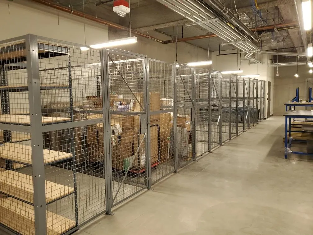 A large storage area with metal wire cages houses various boxes and packages. The room features concrete floors, overhead fluorescent lighting, and a sturdy pallet rack next to empty metal shelves on the left. A blue workbench is situated to the right.
