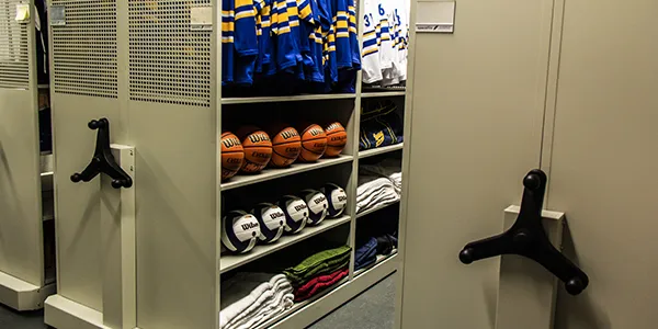 Storage room with spacefile mobile shelving, holding sports equipment. Blue and yellow jerseys hang on top. Basketballs are neatly stacked in the middle section, volleyballs below, and towels folded on the bottom shelf. Handles for moving shelves enhance organization efficiency.