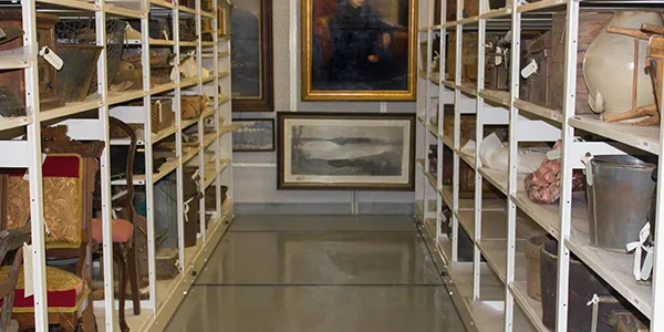A storage room with paintings and various artifacts neatly arranged on spacefile mobile shelving. The organized space features framed artwork on the back wall and tagged items on the shelves. The polished concrete floor enhances the rooms tidy, well-maintained appearance.