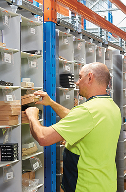 A man in a light green shirt meticulously organizes items on a set of shelves in the storeroom, embodying the efficiency of the Storeganizer system. He reaches for a small box amid other neatly arranged packages, showcasing Pick 360 precision in an industrial storage setting.