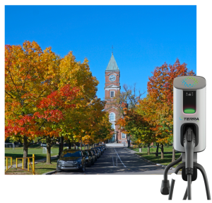 An autumn scene unfolds with colorful trees lining a street leading to a building with a clock tower. Cars, including modern electric vehicles, are parked along the road. In the foreground, an EV charging station stands near what looks like Utah lockers by the sidewalk.