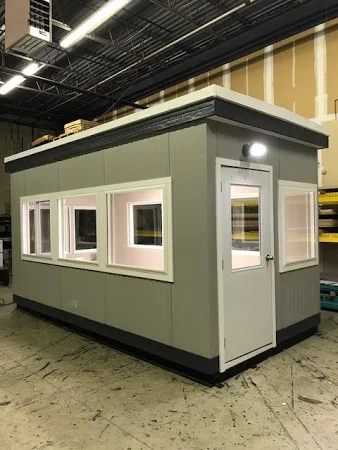 A small, rectangular Utah guard booth with gray walls and a flat roof sits inside a warehouse. It features multiple windows and a white door, surrounded by visible ceiling lights and equipment.