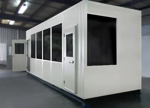 A large white modular office building with multiple windows and a door, designed for shipping and receiving, is set indoors. It sits on a gray platform within a spacious industrial setting, under a ceiling with visible beams and fluorescent lights.