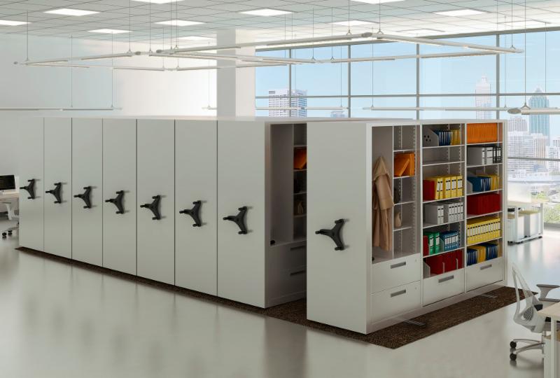 A modern office space features several white, mobile shelving units for efficient storage. One unit is open, displaying organized binders, jackets, and office supplies. Large windows showcase a cityscape in the background, enhanced by bright lighting and minimalistic design.