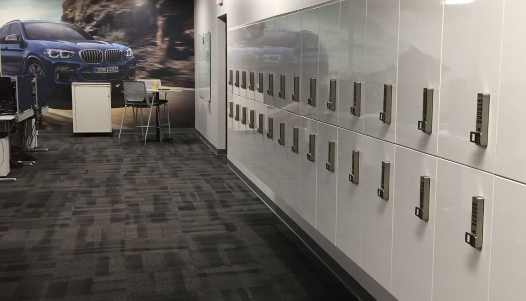The hallway features a row of white lockers with secure storage options, including gray number pads. To the left, a table with two chairs and a computer sits comfortably. A large mural of a blue car on a rugged landscape adorns the back wall.