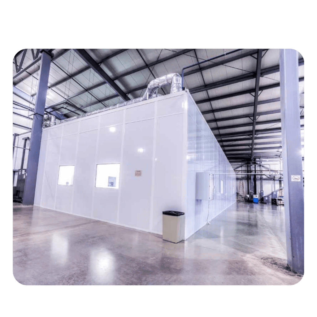 The interior of this spacious industrial building showcases a large, white, panel-built structure with windows. The facility boasts a high ceiling with visible beams and an empty concrete floor. A trash can is discreetly placed beside the structure.
