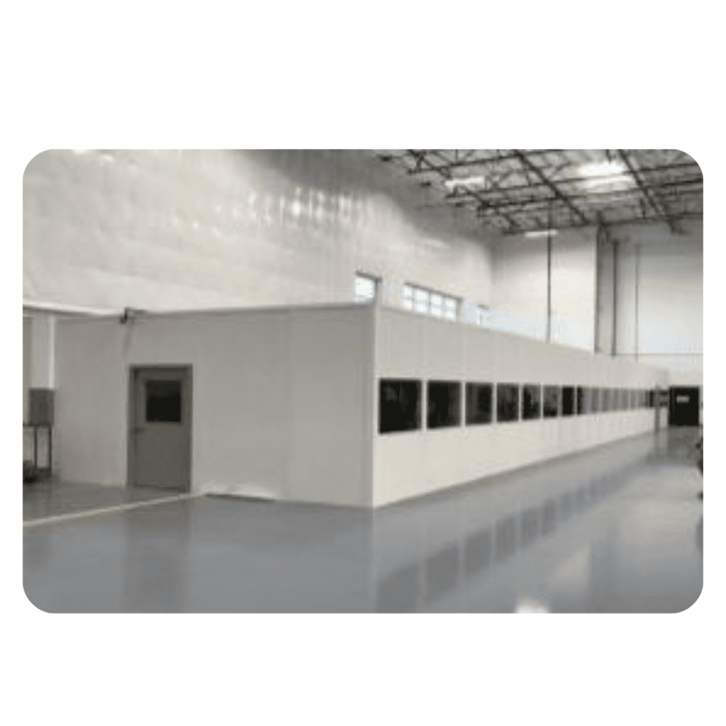 The interior of the large industrial warehouse reveals a long, white partitioned room, cleverly panel built with windows and a door. The space boasts high ceilings and bright, polished floors.