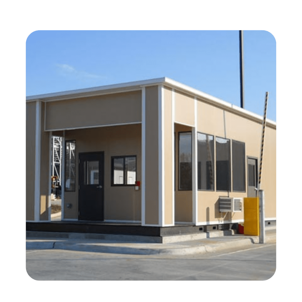A small beige security booth, panel built with large windows and a black door, stands on a paved area. Surrounded by a metal barrier and equipped with an air conditioning unit on the side, it basks under a clear blue sky.