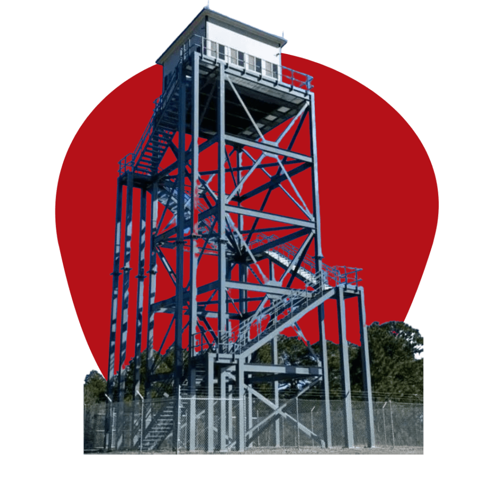 A tall, metal observation tower, reminiscent of an operator cab on a 70-foot structure for the US Military, features multiple staircases and a small room at the top. It stands against a red background, enclosed by a fence with distant trees visible.