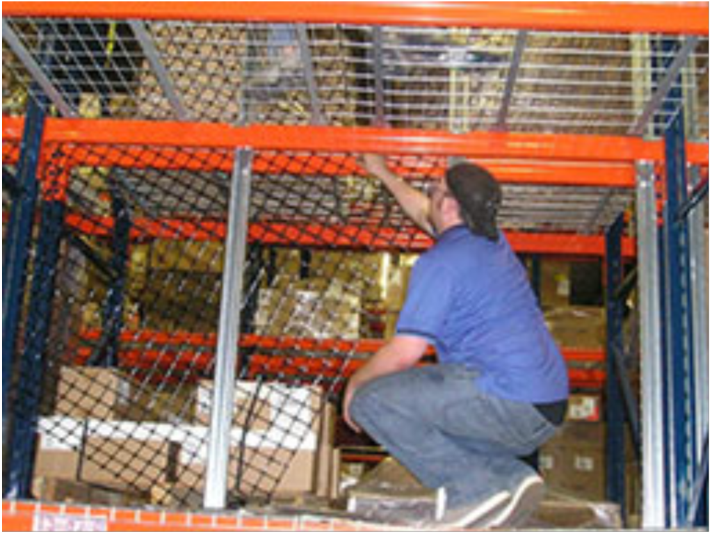 A person in a blue shirt is crouching on a lower shelf of an industrial storage rack, reaching up to organize or retrieve boxes stacked above. The sturdy shelves are secured with pallet rack safety netting for added protection.