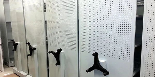 Spacefile mobile shelving units with perforated panels and large rotating handles are shown. The shelves are aligned in a row in a well-lit room, designed for efficient storage and easy access.