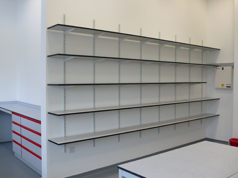 The minimalist setting features an empty white room with wall-mounted shelves in a grid pattern. Red and white cabinets add a touch of color on the left, enhancing the light gray floor and plain white walls—an ideal canvas for comprehensive lab renovation services.