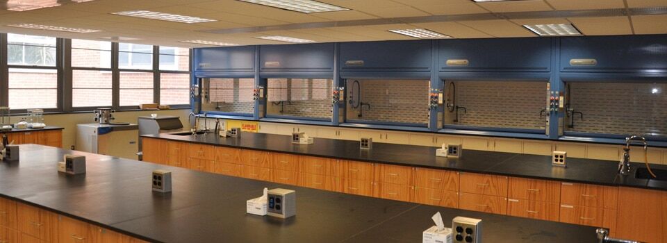 A spacious chemistry laboratory featuring mobile lab tables with adjustable height, long black countertops, multiple wooden cabinets, and blue fume hoods along the back wall. The room is well-lit with fluorescent lights and large windows. Several sinks and electrical outlets are on the counters.