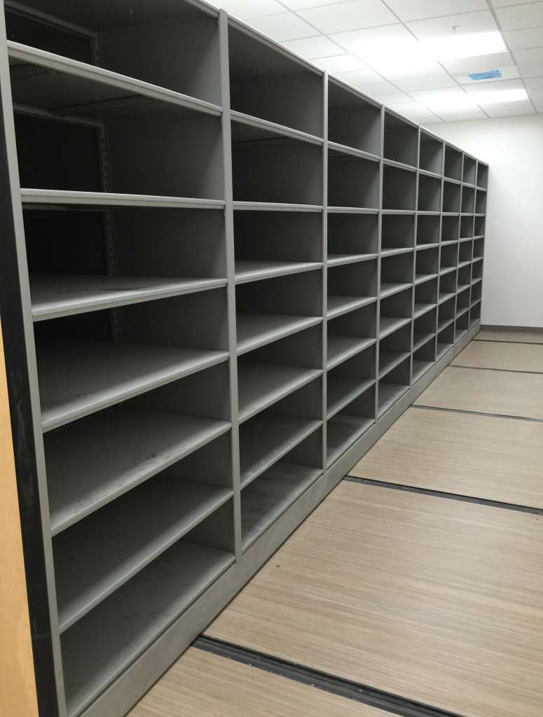 A long, empty storage shelf unit with multiple rows and columns stands against a light-colored wall. The wood-paneled floor reflects the bright overhead lights, revealing the potential of customizable mobile shelving units for lab equipment to transform and maximize laboratory space efficiently.