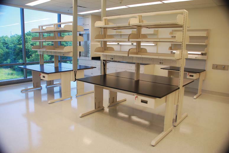 A bright, spacious laboratory with large windows displaying a greenery view features ADA-compliant black-topped workbenches and white shelves above them. The room is empty, yet its clean and organized appearance speaks to the vocational high schools dedication to optimizing lab spaces.