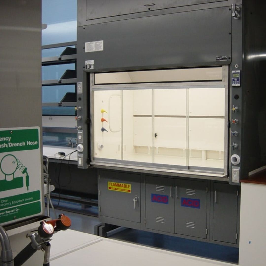 A laboratory fume hood with a glass front, featuring several knobs and warning labels, exemplifies premium laboratory safety solutions. A green emergency wash/drench hose sign stands in the foreground amid well-organized equipment, highlighting a workspace optimized for safety and efficiency.