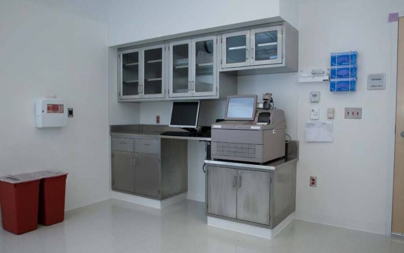 The laboratory room, perfect for quality control in distilleries, features metal cabinets, a computer, and a printer on the counter. The walls are pristine white with a red recycling bin nestled in the corner, complemented by a wall-mounted fire alarm and numerous switches and labels.