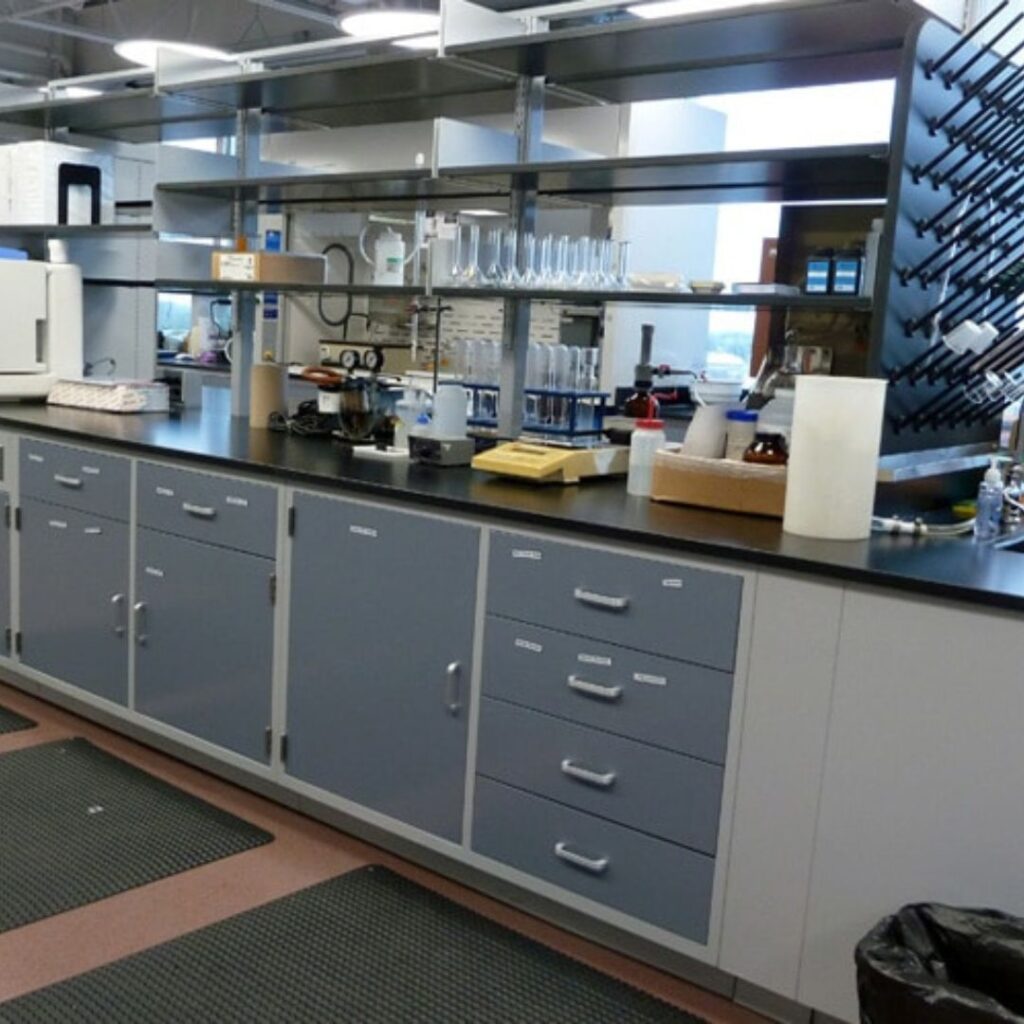 A process research lab featuring a countertop with test tubes, bottles, and equipment. Cabinets and drawers are below, with shelves for additional gear above. Pegboards for drying glassware adorn the wall, showcasing smart distillery laboratory design elements.