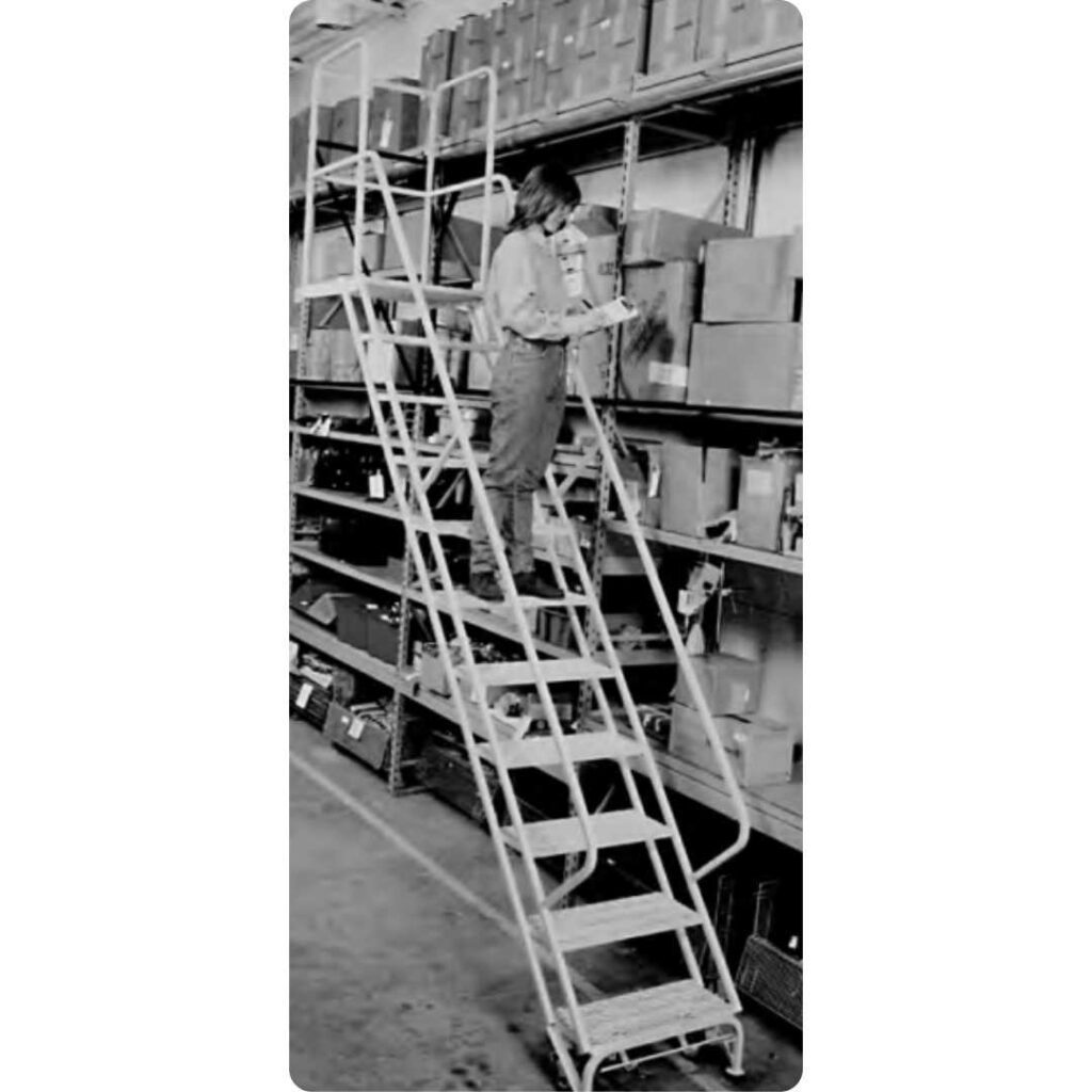 A person stands on a rolling ladder from Cotterman Ladders Company in a warehouse, reaching for items on a high shelf. The shelves are packed with boxes and various items, all captured in striking black and white.
