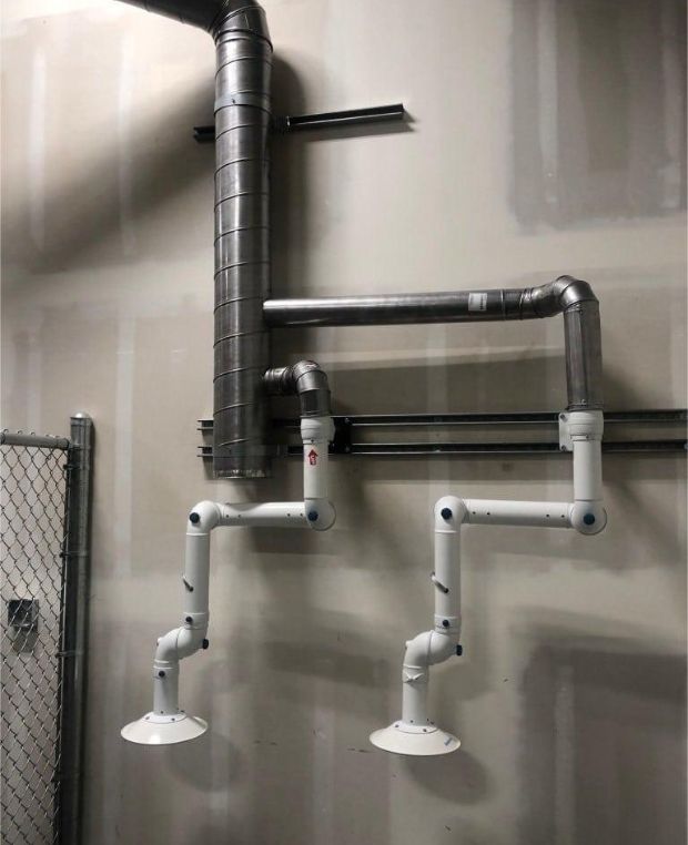 A wall-mounted advanced laboratory snorkel arm ventilation system features metal ducts and two adjustable white exhaust arms. The setup resembles the shape of a stick figure horse, with a chain-link fence partially visible on the left.