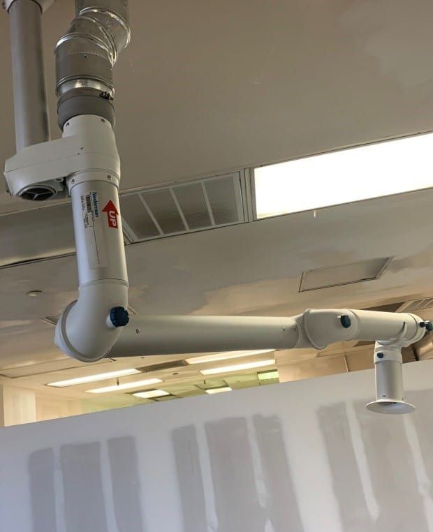 An advanced laboratory snorkel arm ventilation system hangs from the ceiling, featuring adjustable joints for precision. Made of durable white metal, it connects seamlessly to an overhead duct. The scene is set against a backdrop of fluorescent-lit ceiling tiles.