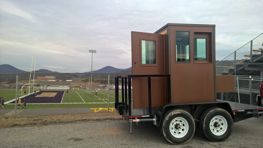 A versatile ticket booth, small and brown with windows, is mounted on a trailer with dual wheels. Its parked by the football field, near metal bleachers. In the backdrop, goalposts stand tall against the majestic mountains.