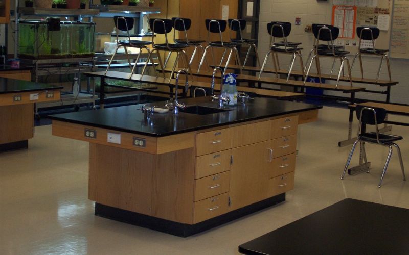 The classroom boasts black-topped lab tables with durable epoxy resin countertops, complemented by wooden cabinets and chairs stacked on benches. It features sinks, a fish tank in the corner, and wall posters. Light-colored tile covers the floor, enhancing an adaptable and efficient learning space.